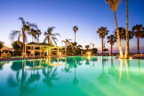 Piscina con palmeras y una zona cubierta, rodeada de follaje.