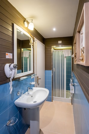 Un baño con lavamanos blanco, secador de pelo, cabina de ducha de vidrio y espejo con iluminación.