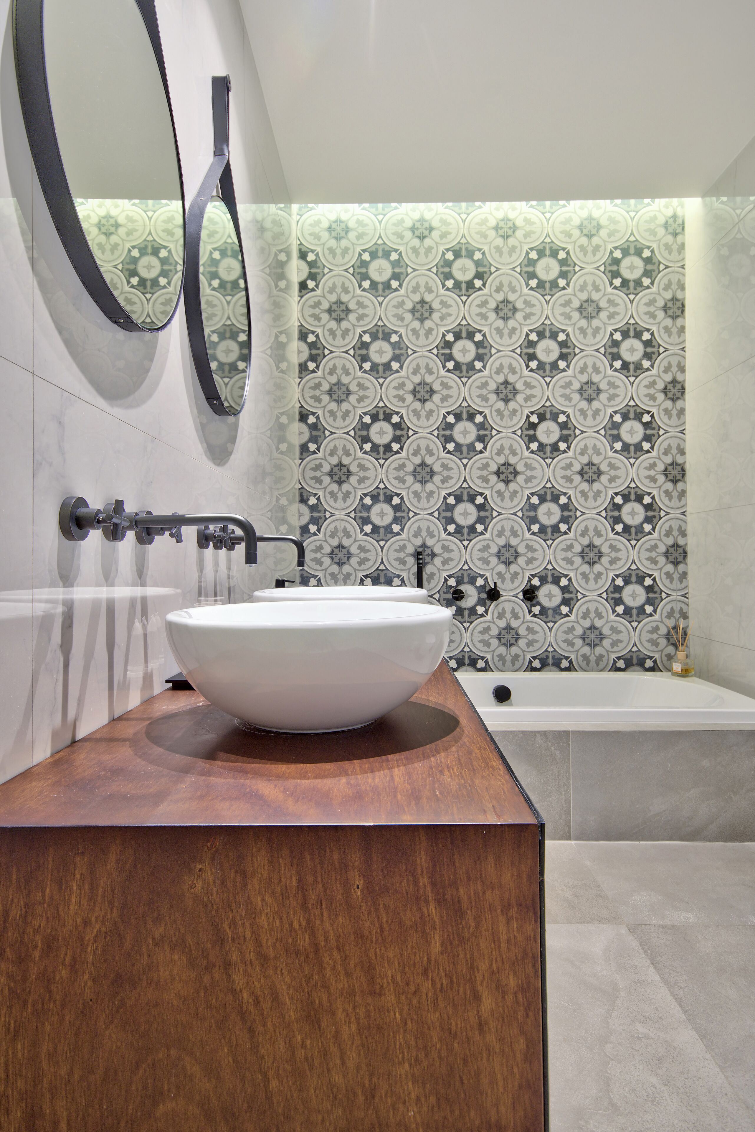 Une salle de bain avec un lavabo sur colonne blanc, un meuble-lavabo en bois, une baignoire et un dosseret de carrelage à motifs.