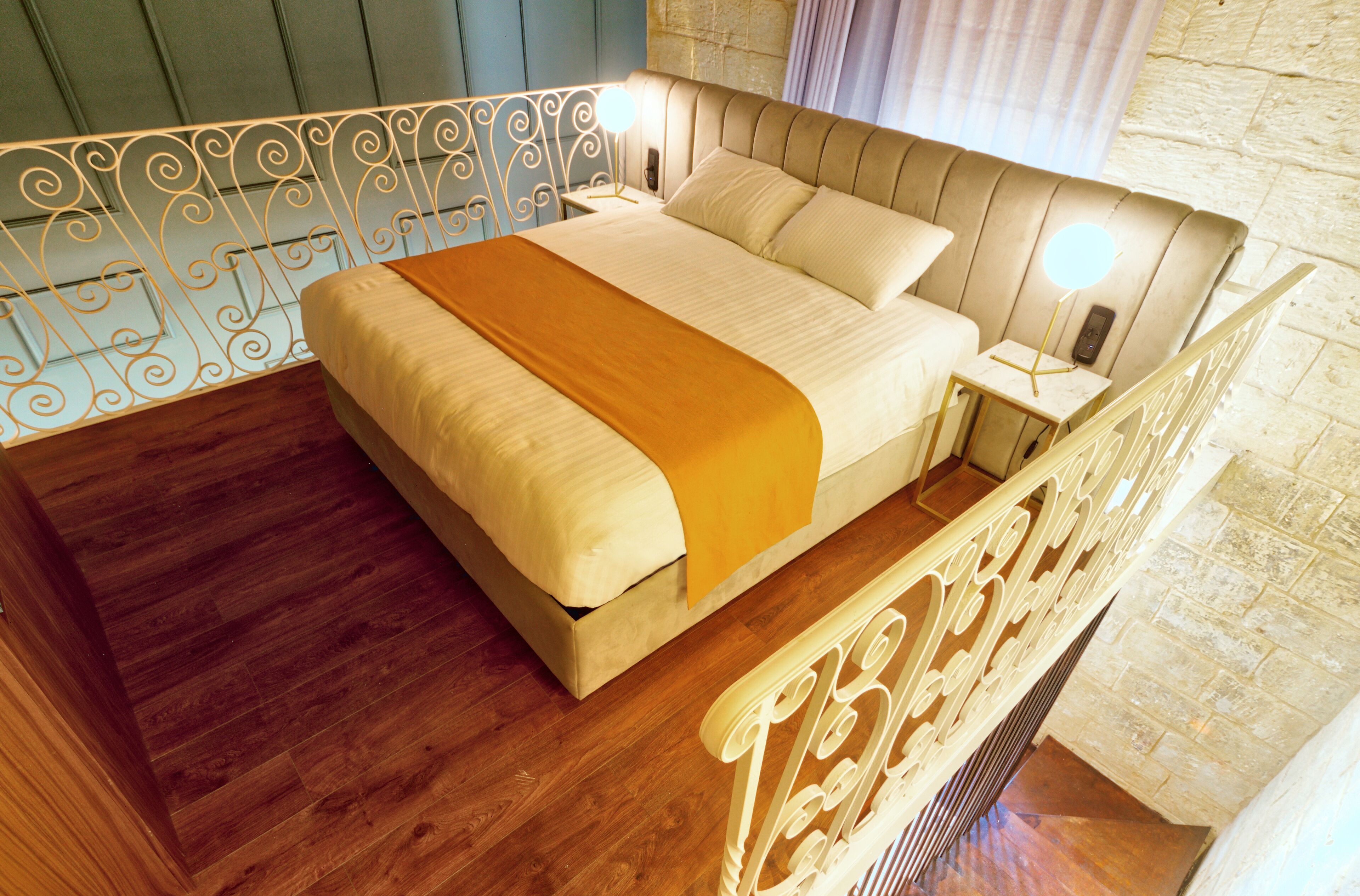 A bedroom with a wooden floor, a bed with a beige bedspread and a yellow throw, a nightstand with a lamp, and a stone wall.
