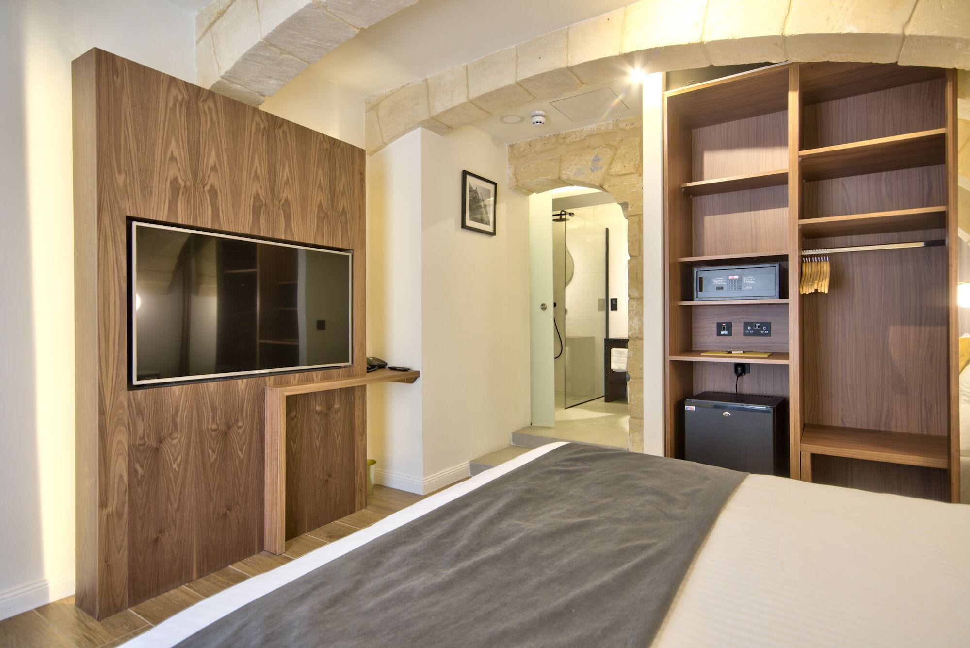 Un dormitorio con una TV montada en un panel de madera, un vestidor con estantes y una cama con una manta gris.