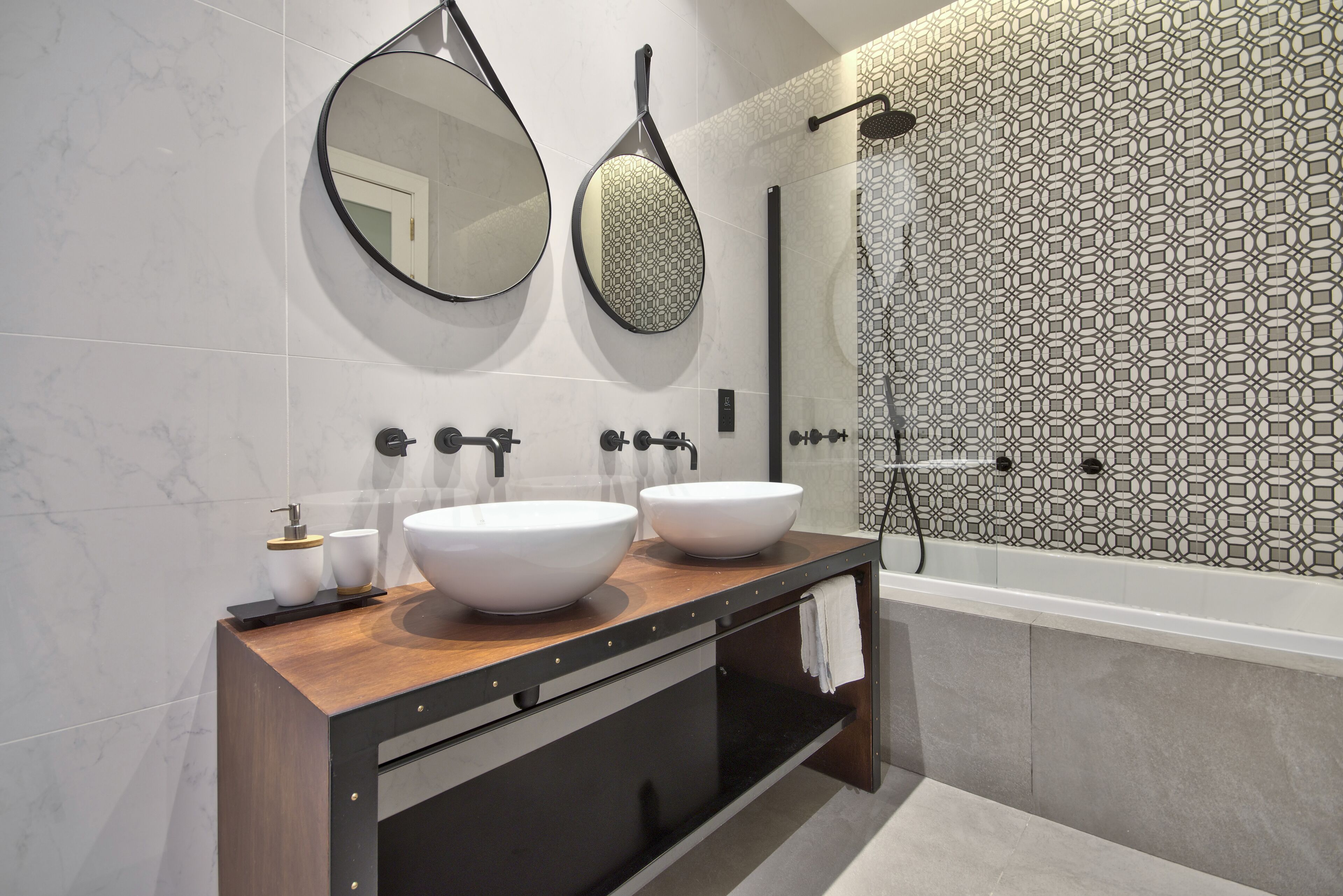 A modern bathroom with two large white sinks, a wooden vanity, a round mirror, and a patterned shower wall.