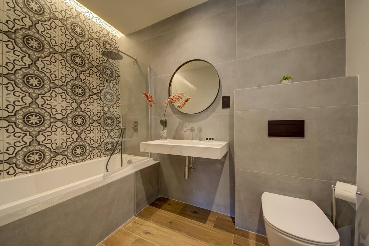 A modern bathroom with a patterned tile wall, a white sink, a round mirror, and a toilet.