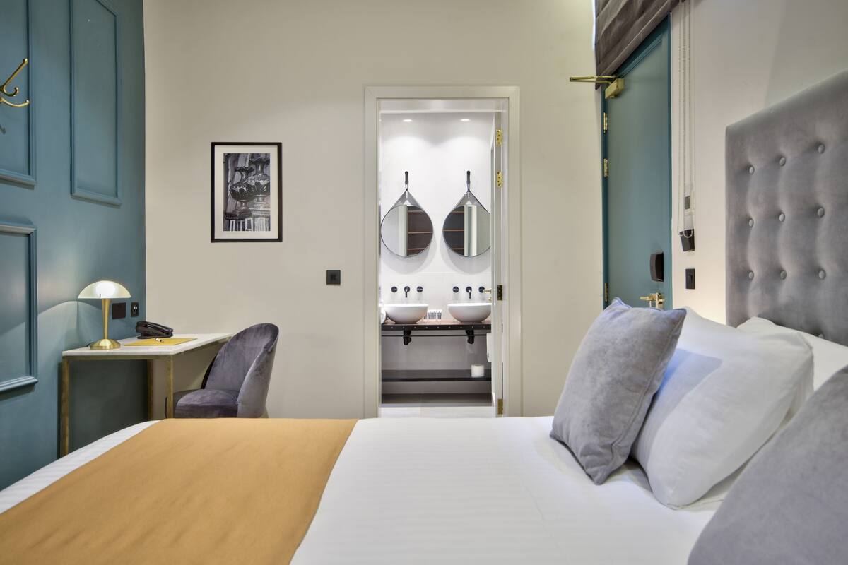 A hotel room with a bed, a desk with a lamp, a chair, a bathroom with a sink and mirror, and a door with a handle.