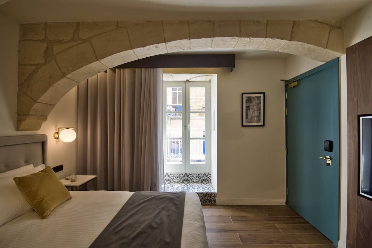 A bedroom with a stone arch, a bed with a headboard, a nightstand, a door, and a view through a window.