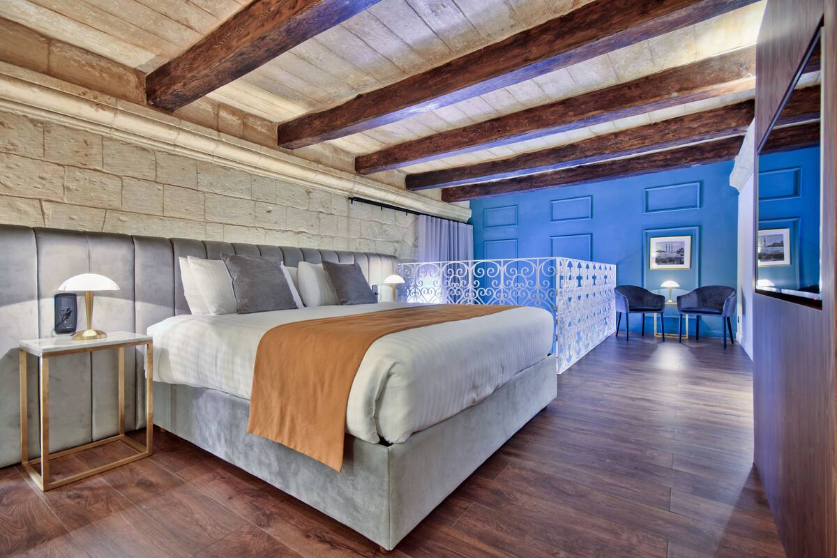 A modern bedroom with a large bed, wooden beams, a brick wall, a blue accent wall, and a small seating area.