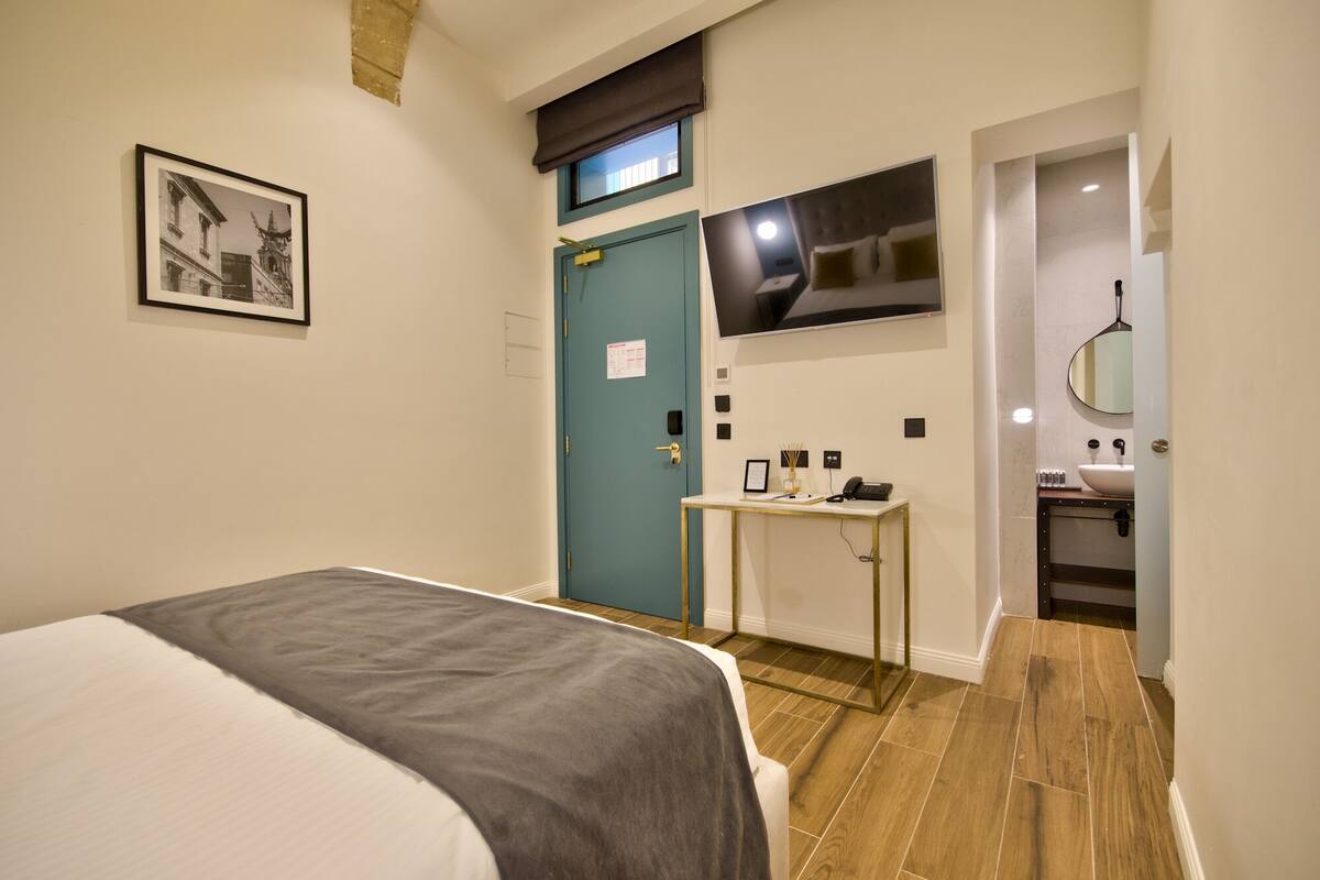 A hotel room with a bed, a desk with a computer, a TV mounted above the desk, and a bathroom with a sink and mirror.