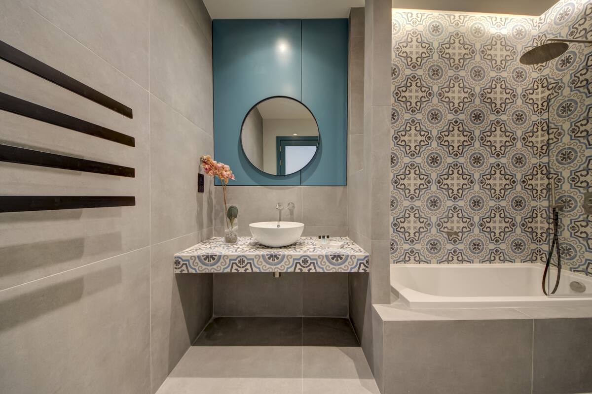 A modern bathroom with a large bathtub, a round mirror, and a patterned tile backsplash.