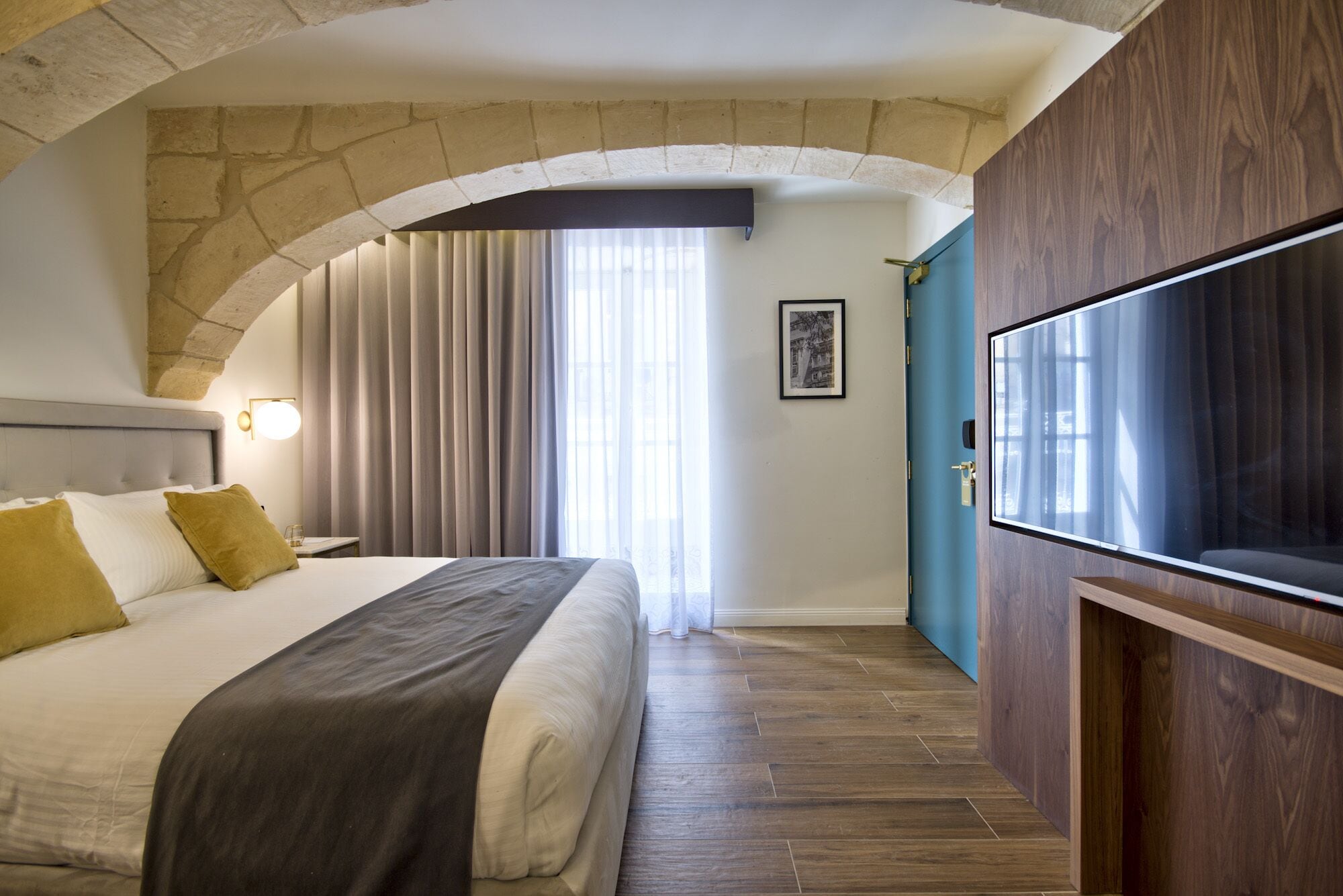 A hotel room with a stone arch ceiling, a large bed with a grey headboard, a wooden TV stand, and a large window with curtains.
