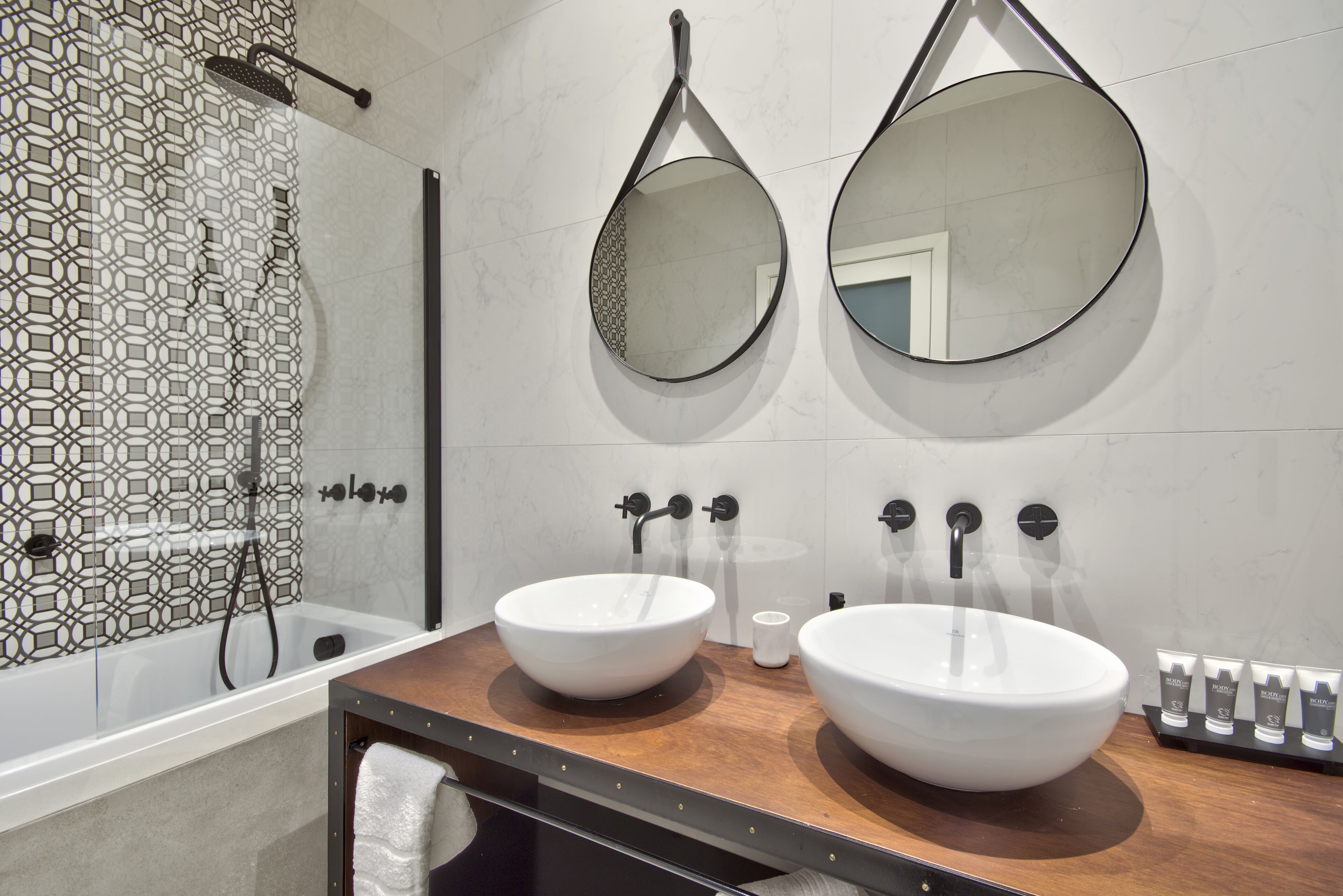 A modern bathroom with two large white sinks, a wooden vanity, a large mirror, and a shower with a geometric tile design.