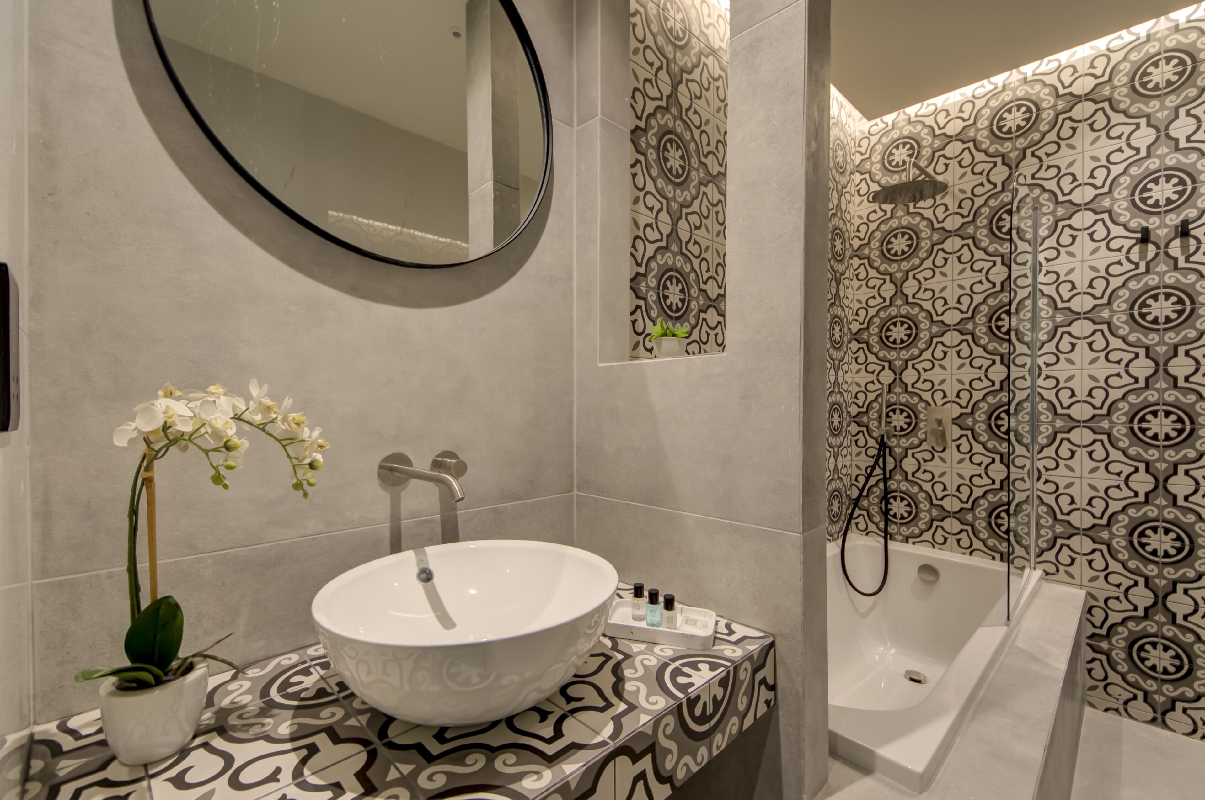 Une salle de bain avec un lavabo blanc, un miroir, une orchidée en pot et un mur de douche orné d’un motif.