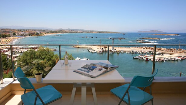 Ein Balkon mit Tisch, zwei Stühlen, einem Blumentopf und einer Zeitschrift. Der Blick umfasst einen Hafen mit Booten, einen Strand und ferne Berge.