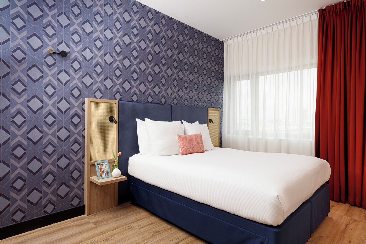 A neatly made bed with white and pink pillows, a wooden nightstand with a picture frame and a vase, and a patterned wall.