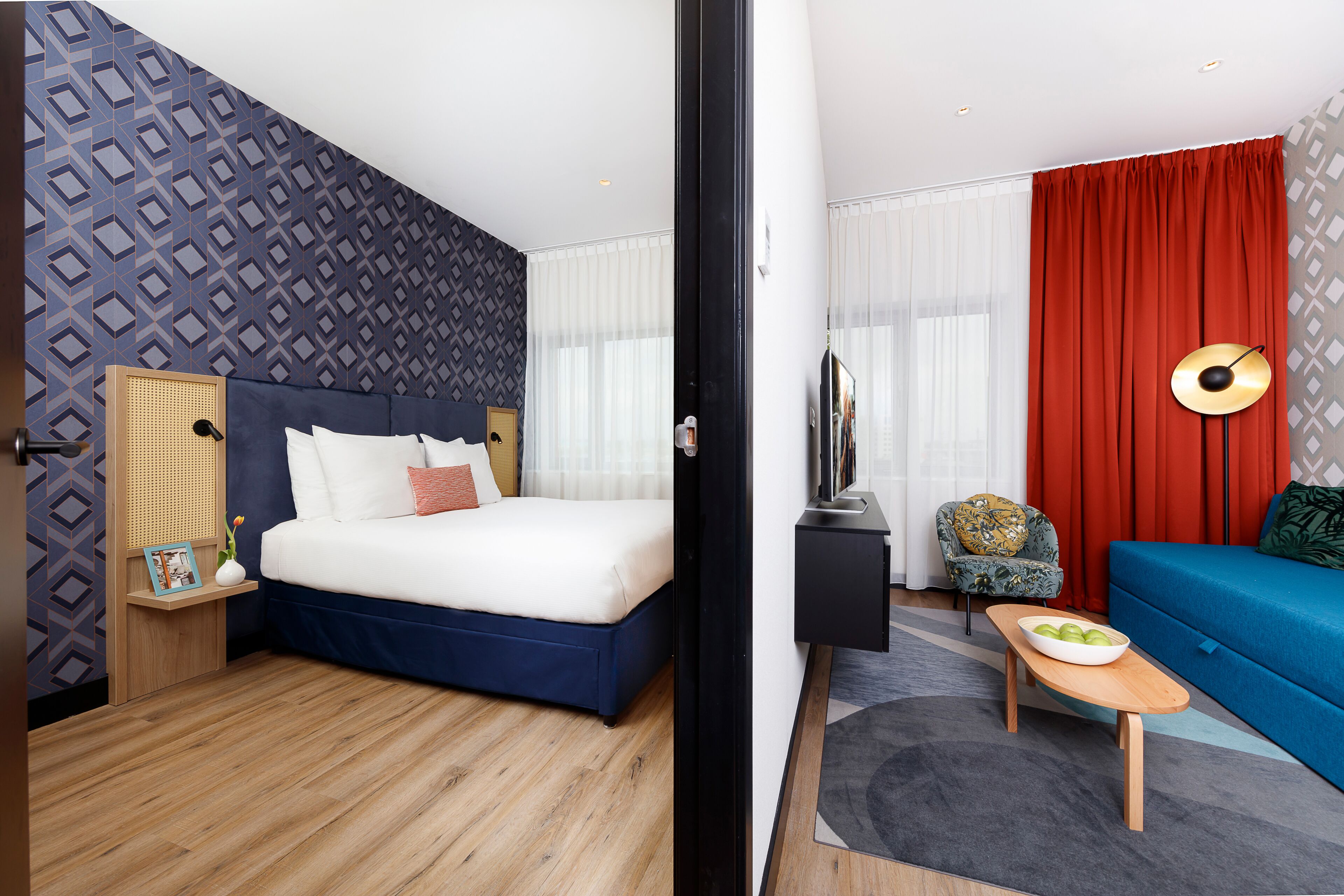 A hotel room with a bed, a sofa, a TV, a small table with a bowl, and a chair.