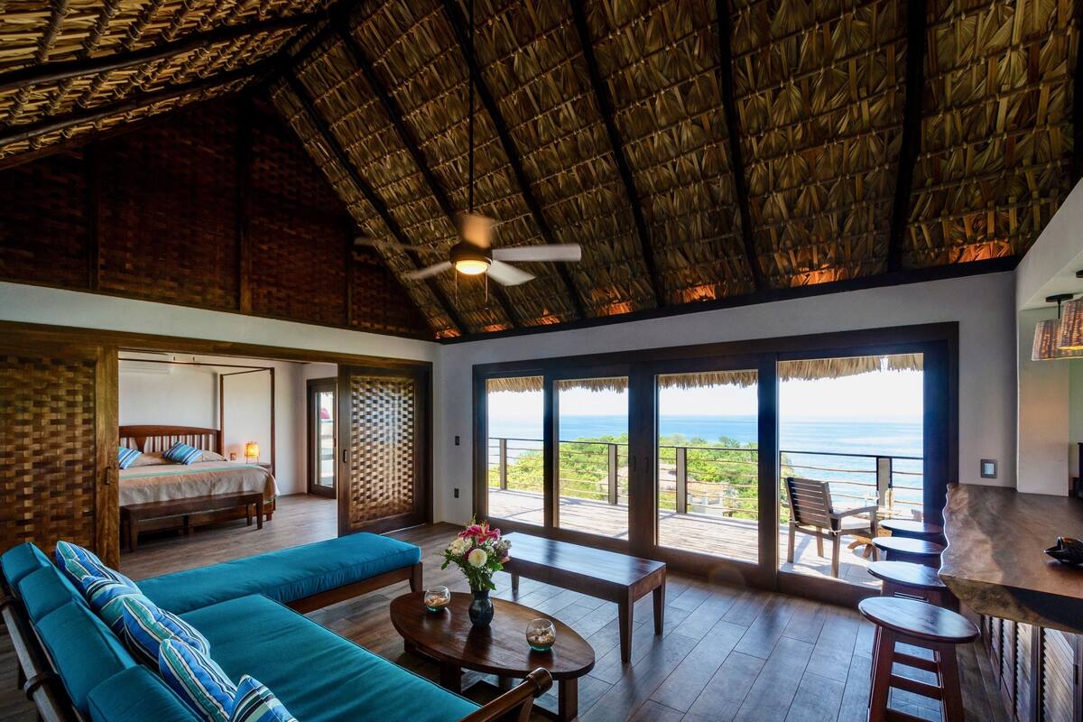 A spacious room with a large bed, a sofa, and a dining area. There is a view of the ocean through large windows.
