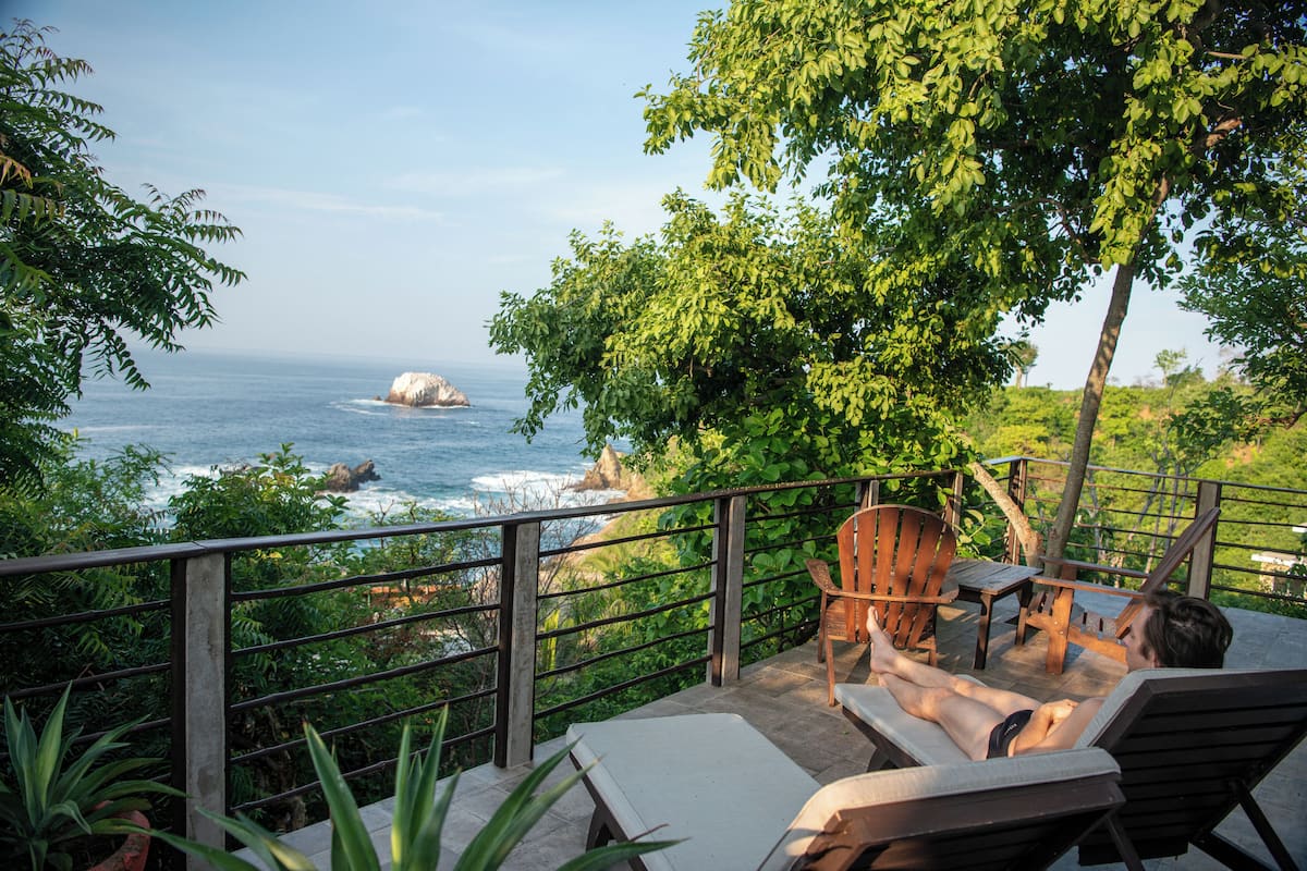 A balcony with a view of the ocean, wooden chairs, and a table.