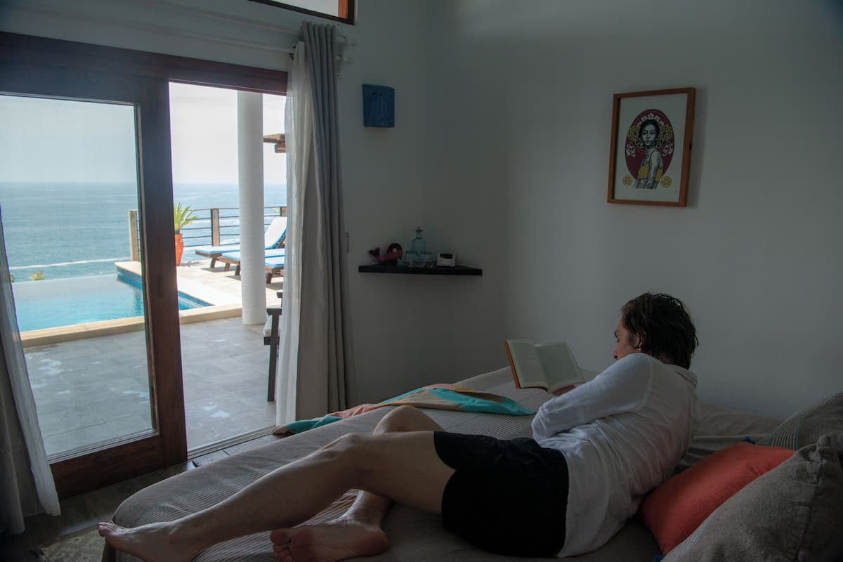 A room with a bed, a window with a view of the ocean, a small shelf with decorative items, and a framed picture on the wall.