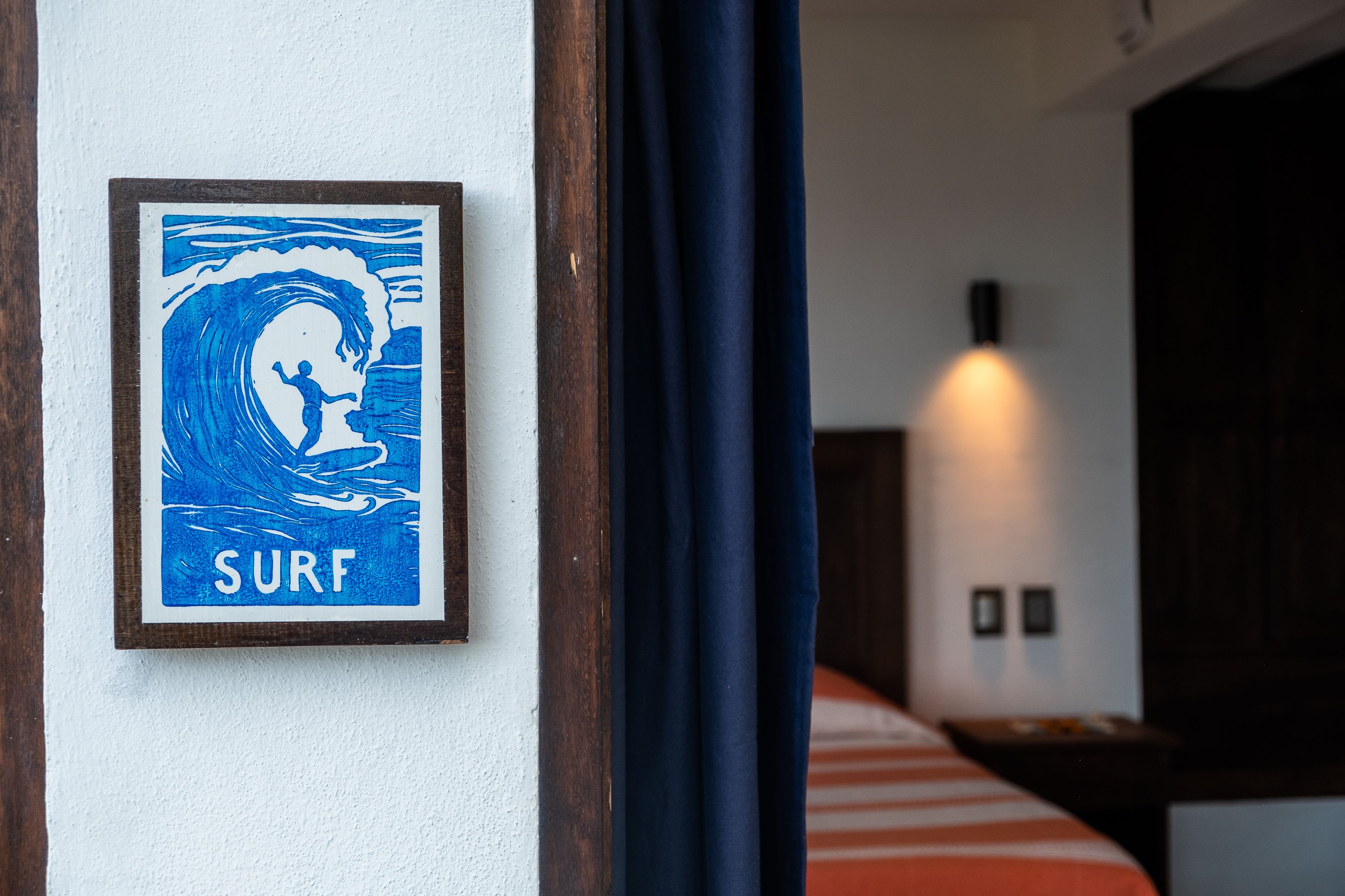 Une œuvre d’art encadrée sur le thème du surf est accrochée au mur, un lit est recouvert d’une couette à rayures, et une table de chevet est agrémentée d’une lampe.
