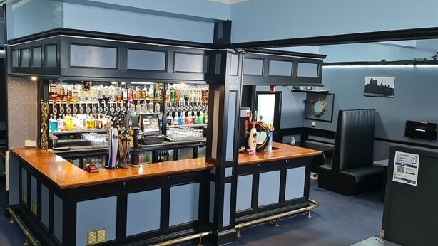Zona de bar amb barra, amples prestatges plens de garrafes de licors i una caixa registradora.