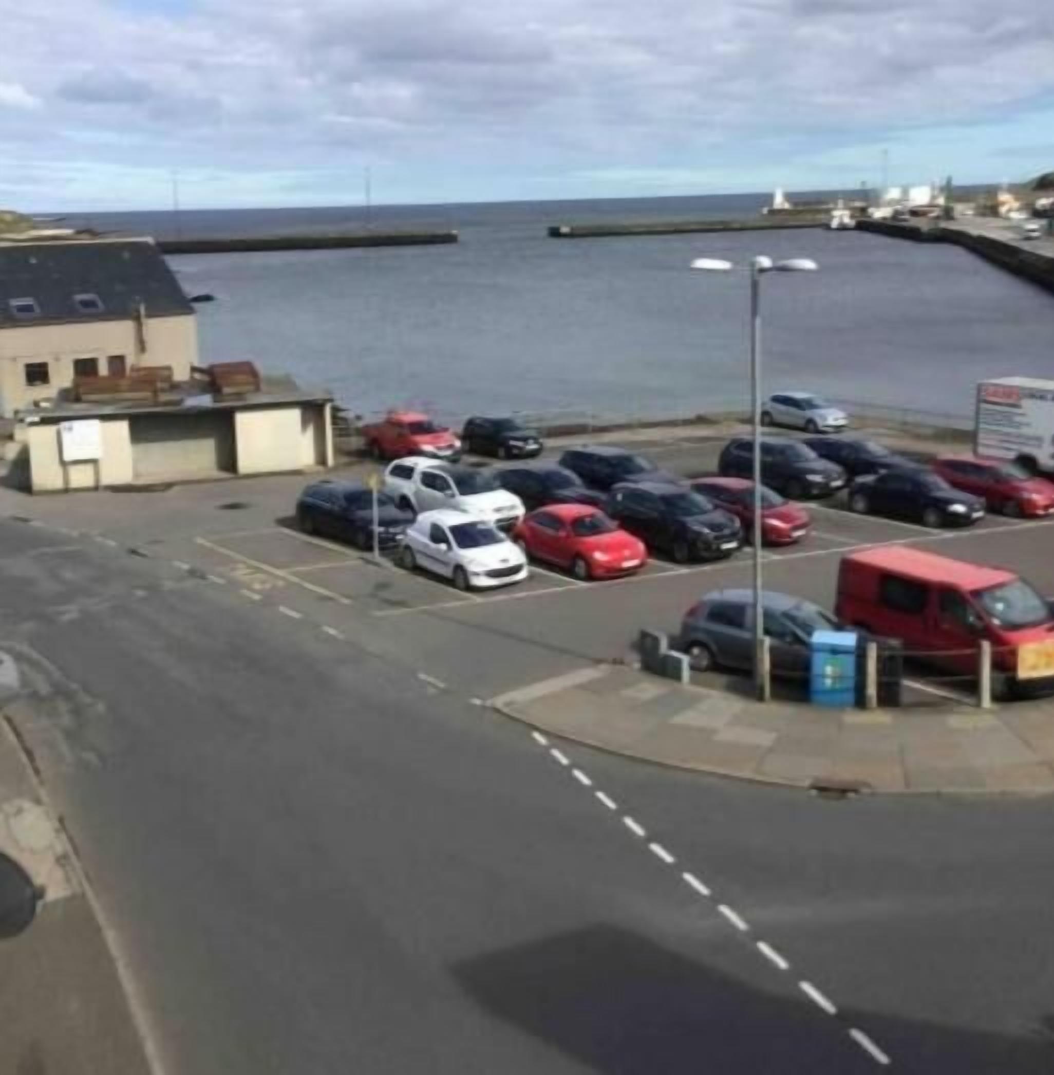 A parking lot with cars and a red van, a building with a sloped roof, and a body of water in the background.