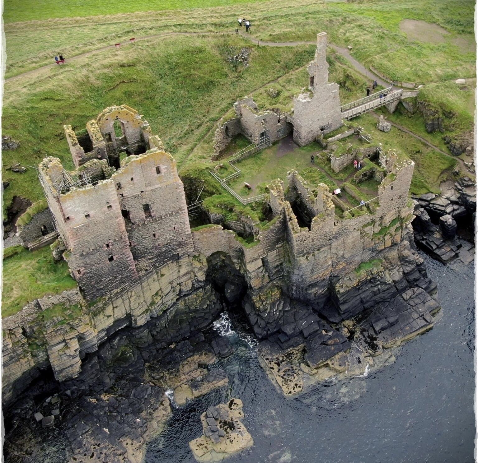 An aerial view of a historic castle complex with multiple towers and ruins, situated on a grassy cliff overlooking the sea.