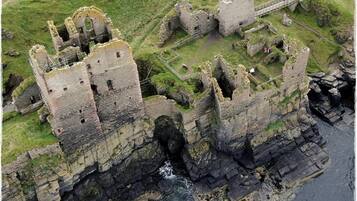 An aerial view of a historic castle complex with multiple towers and ruins, situated on a grassy cliff overlooking the sea.