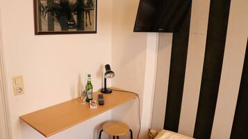 A small room with a wooden desk, a chair, a bed, a TV mounted on the wall, and a framed picture above the desk.