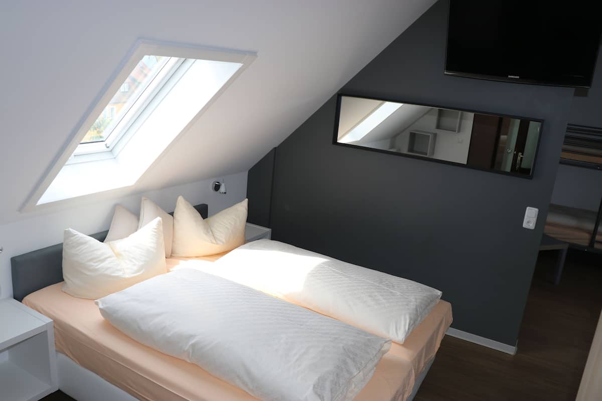 Un dormitorio con tragaluz, una cama con ropa de cama blanca, una mesita de noche, un televisor montado en la pared y un espejo.