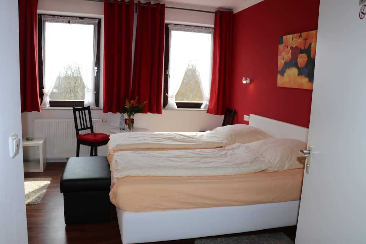 Un dormitorio con dos camas, una silla, una mesita y un ventanal con cortinas rojas.