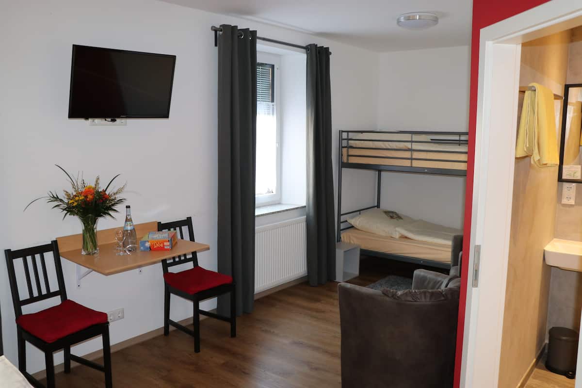 Habitación pequeña con literas, una mesita con sillas, un televisor montado en la pared y una ventana con cortinas.