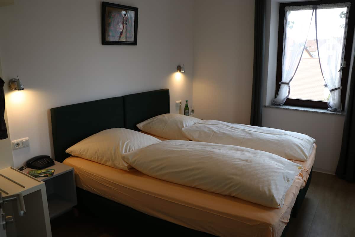Una cama bien tendida con sábanas blancas, una mesita de noche con lámpara, y una ventana con cortinas.