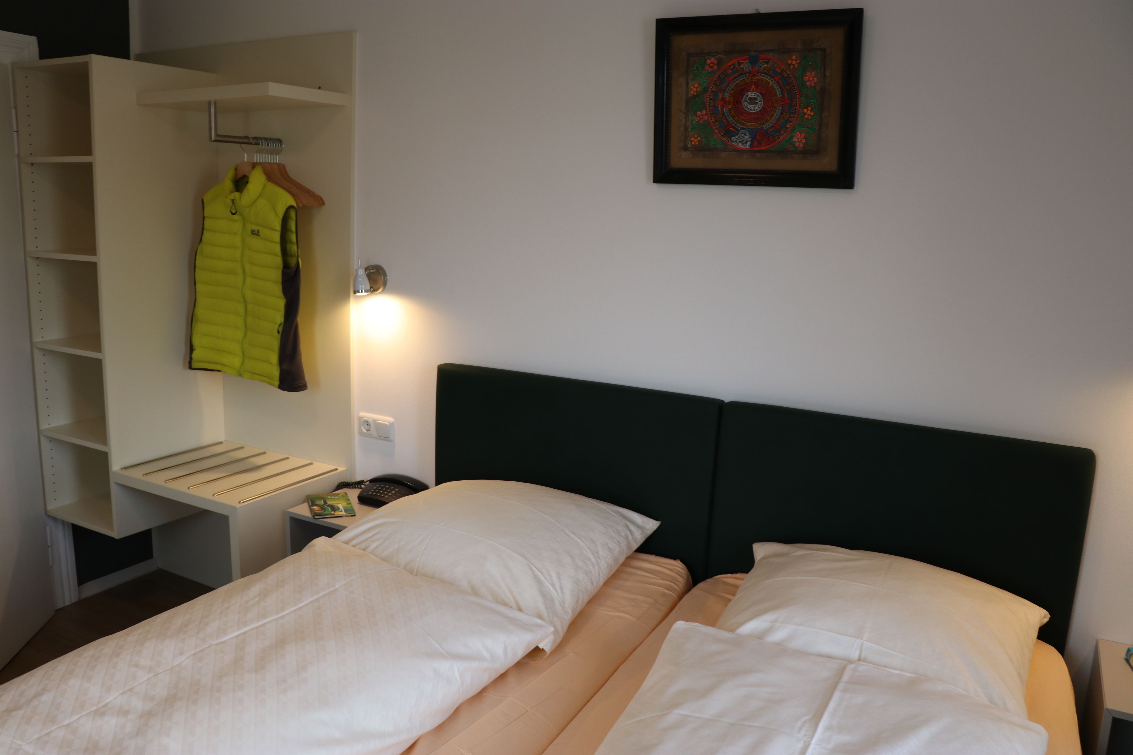 A neatly made bed with white linens, a hanging coat rack with a yellow jacket, and a framed artwork on the wall.