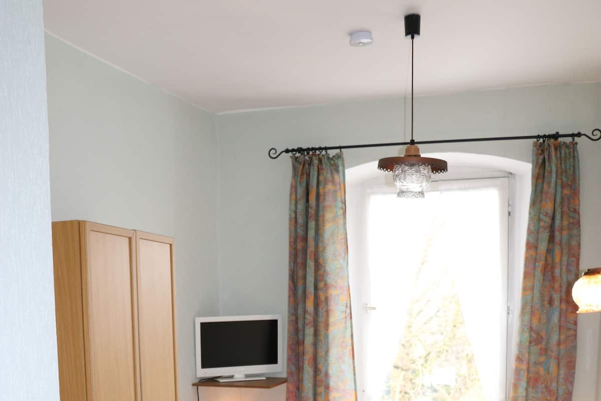 Una habitación pequeña y bien iluminada, con una cama, un escritorio con una computadora y un ventanal con cortinas.
