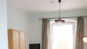A small, well-lit room with a bed, a desk with a computer, and a window with curtains.