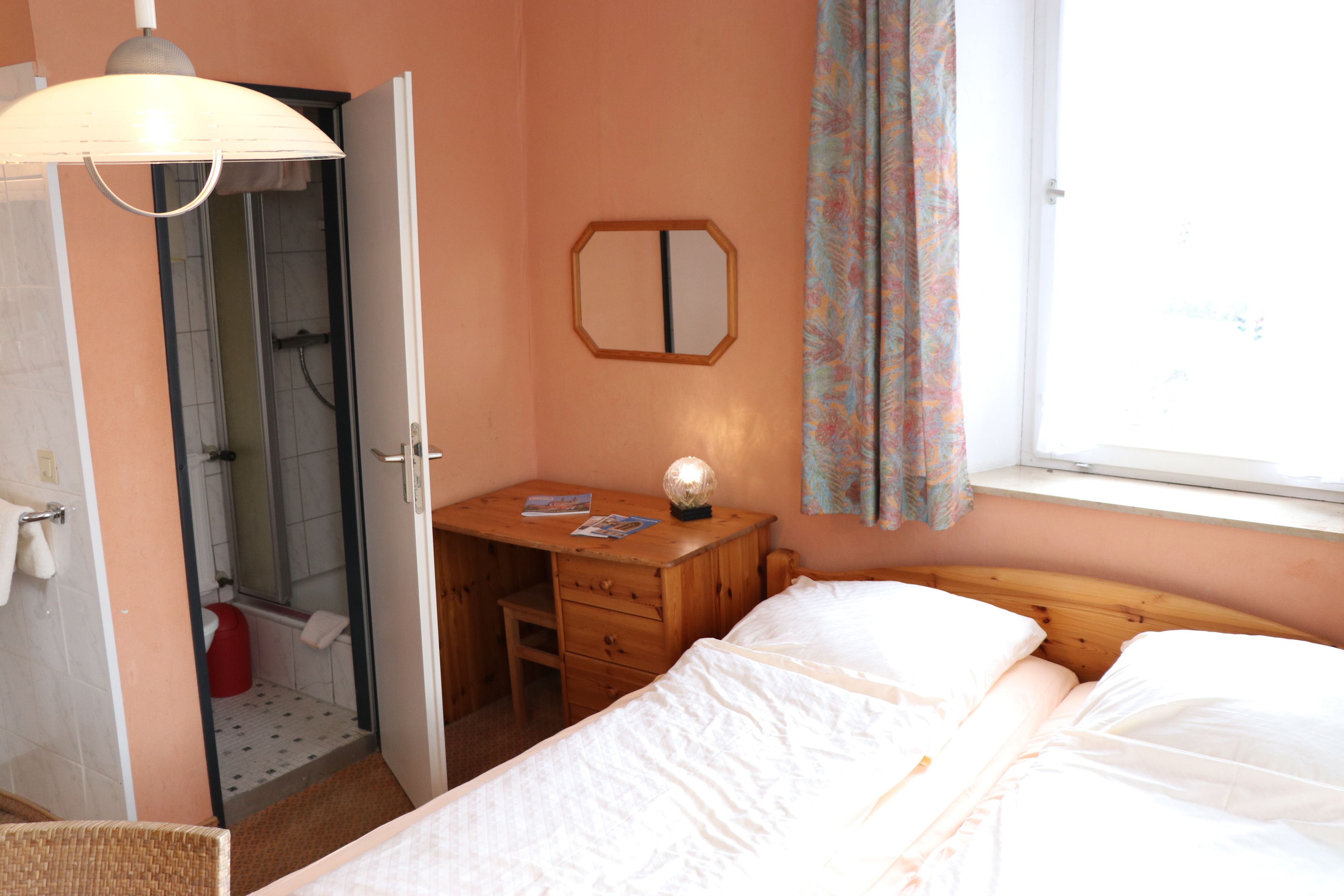 A bedroom with a bed, a wooden desk, a mirror, and a window with curtains.