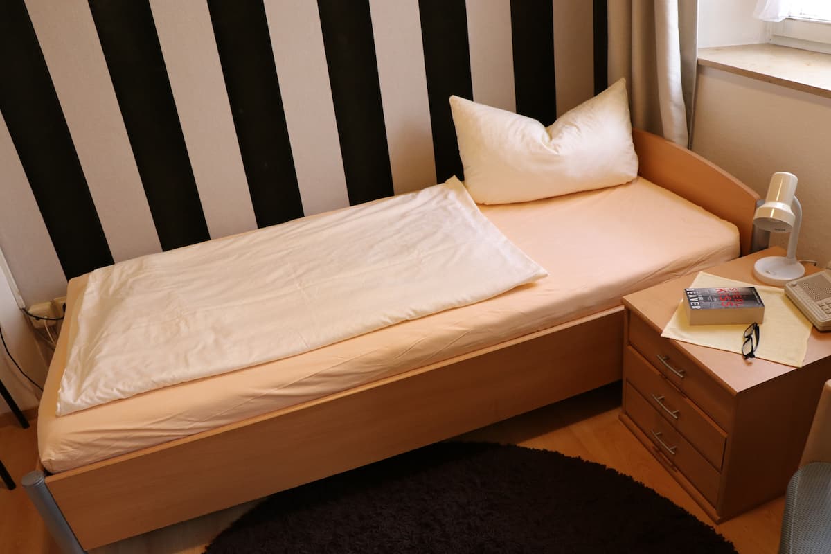 Una cama individual con colchón y almohada blancos, una mesita de noche de madera con una lámpara, un libro y un par de gafas.