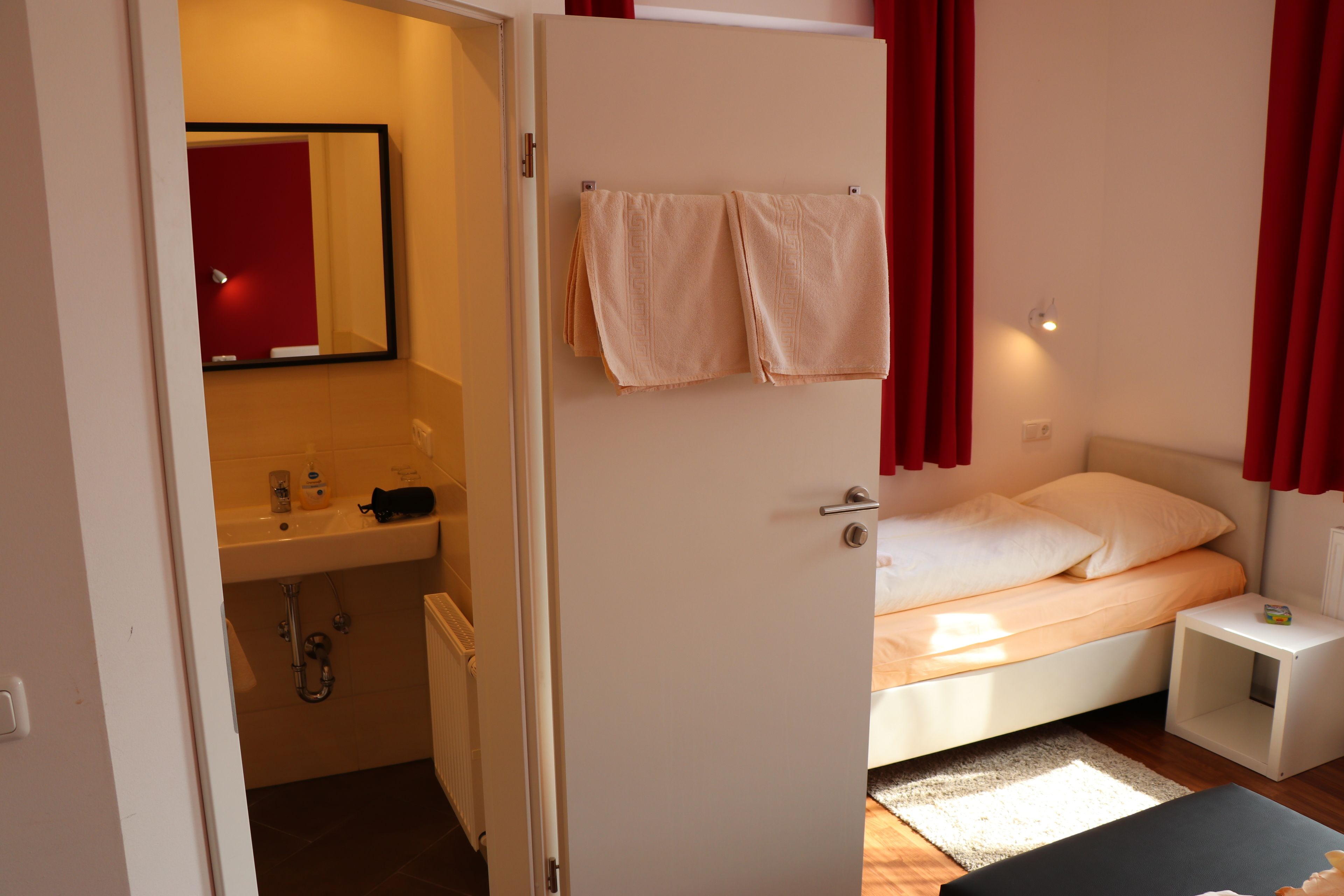 A bedroom with a bed, a nightstand, a small table, a bathroom with a sink and mirror, and red curtains.