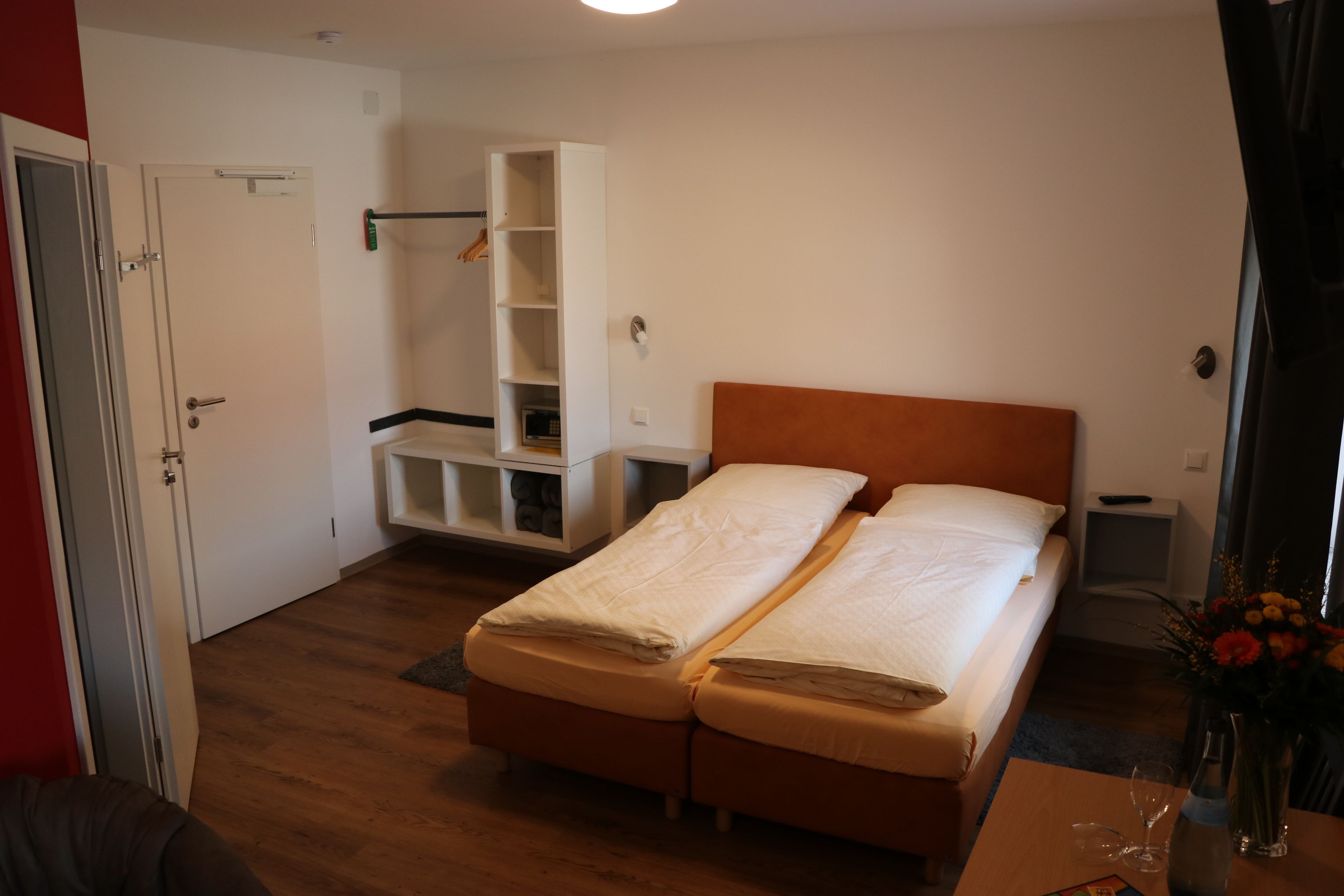 Habitació d'hotel amb dos llits individuals, un terra de fusta, un armari blanc, una taula petita amb un ram i una porta.