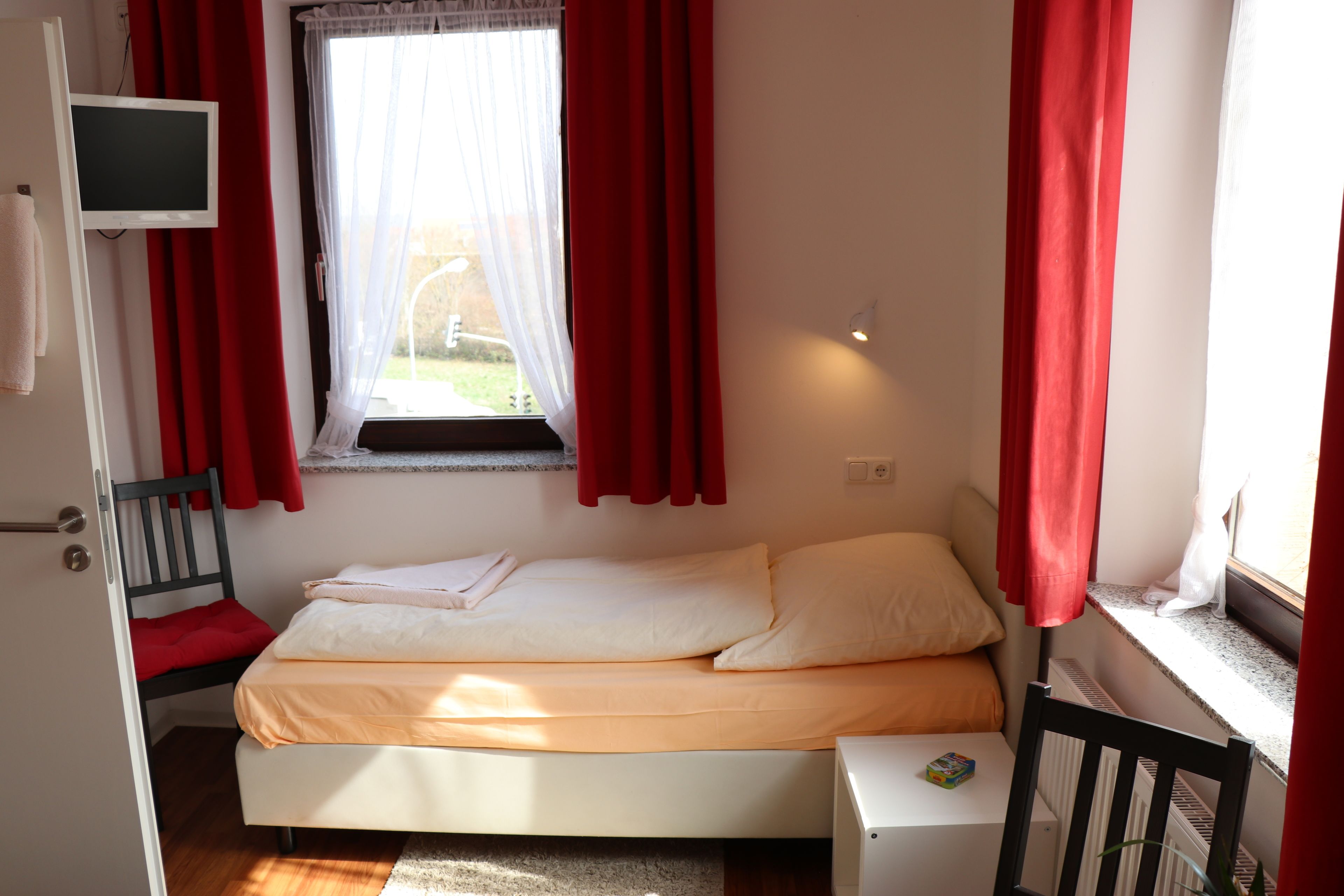 Una cama individual con ropa de cama blanca y naranja, una mesita de noche pequeña, una silla, un televisor montado en la pared y una ventana con cortinas rojas.
