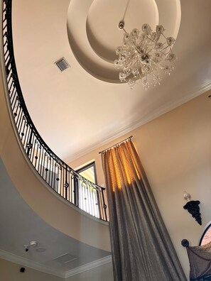 Eine geschwungene Treppe mit schwarzem Geländer, einem Kronleuchter und einem Fenster mit Vorhängen.