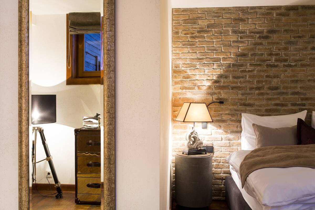 Un dormitorio con pared de ladrillo, una cama con almohadas, una mesita de noche con lámpara y un aparador con cajones.