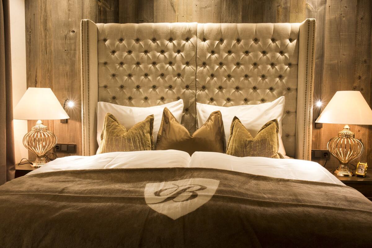 Una cama grande con cabecera capitonada, varios almohadones y una manta con un logotipo. Hay dos lámparas de noche y una mesita con una lámpara encima.