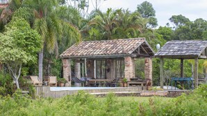 A rustic stone structure with a tiled roof, surrounded by lush greenery and palm trees. - Vieja Hacienda (Imbituba)
