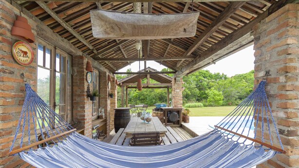 A covered outdoor area with a hammock, wooden table, and brick walls. - Vieja Hacienda (Imbituba)