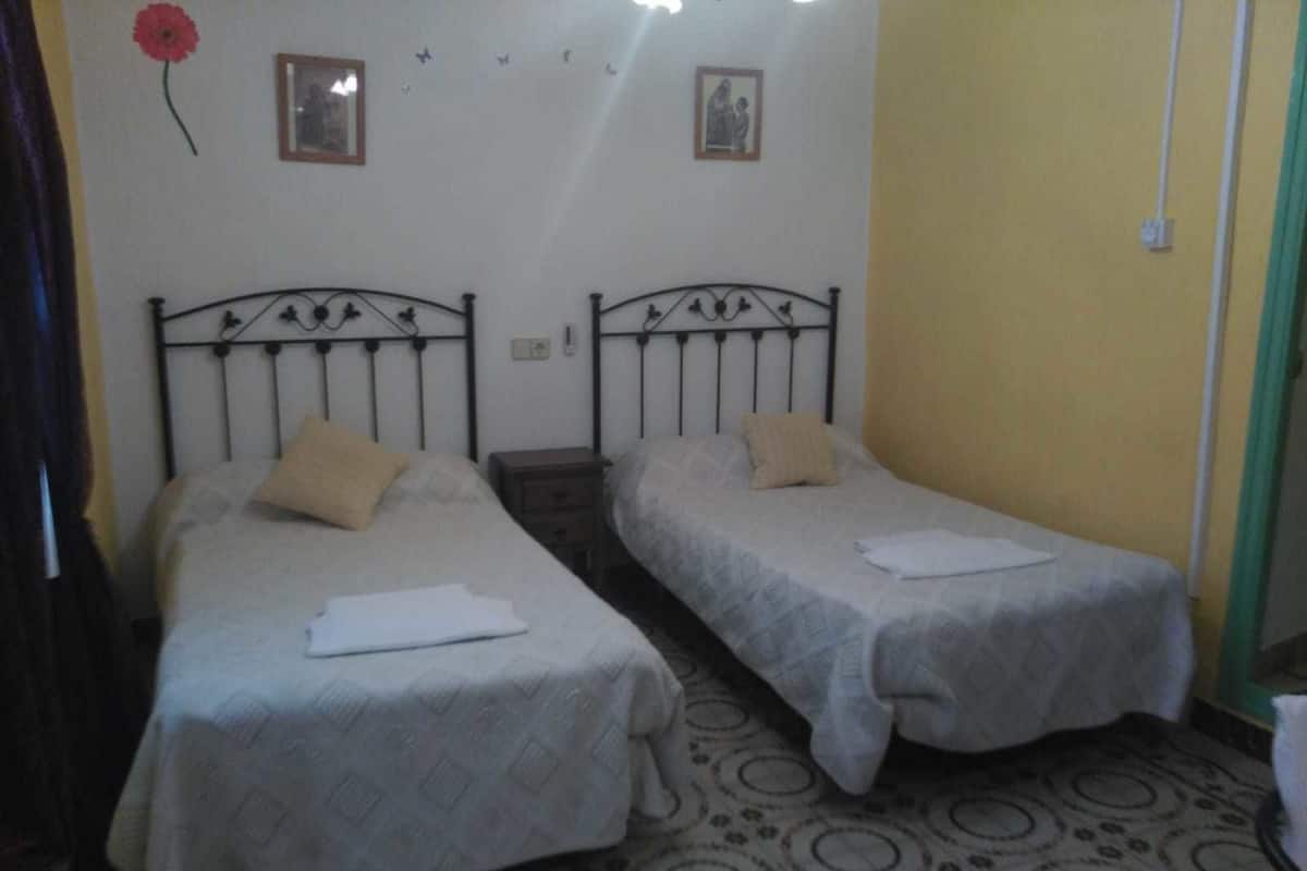 A room with two single beds, each with a pillow and a folded white cloth on the bed. The walls are adorned with framed pictures and a red flower decoration.