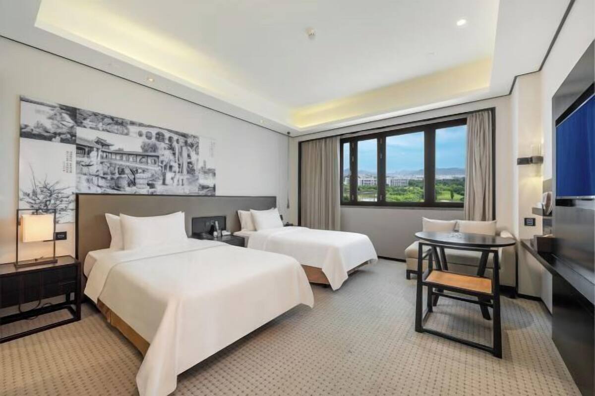 A hotel room with two beds, a desk, a chair, a TV, and a large window with a city view.