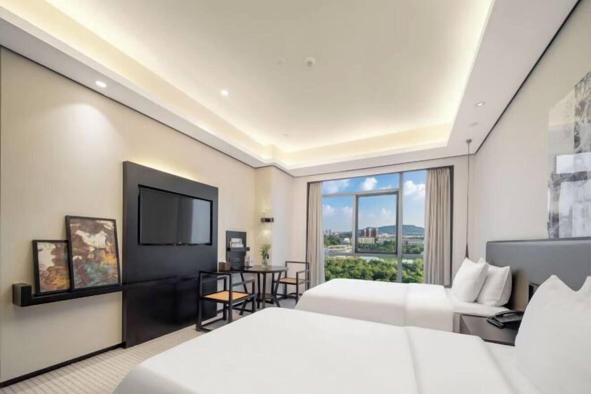 A modern hotel room with a large bed, a desk with a chair, a flat-screen TV mounted on the wall, and a large window offering a city view.