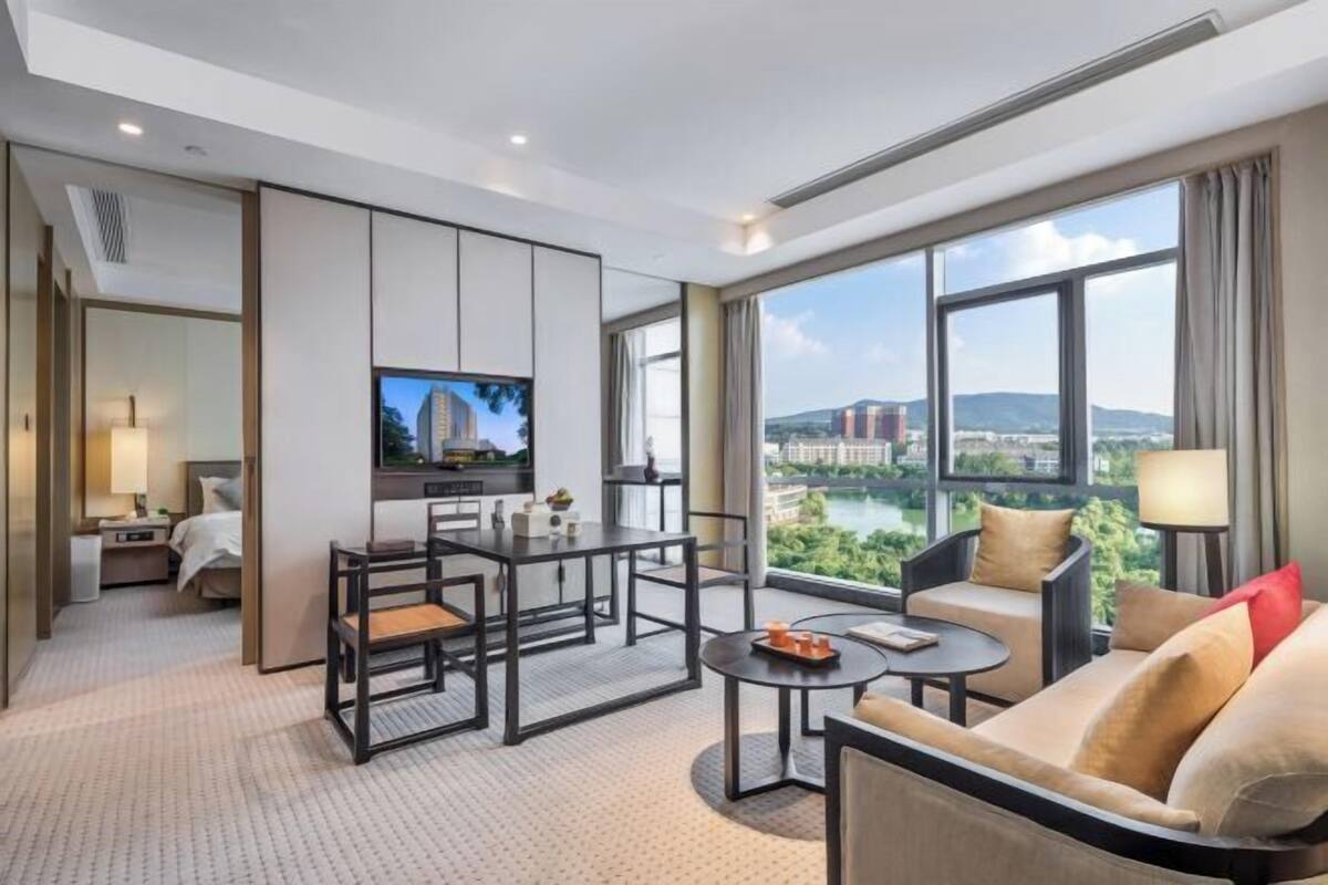 A modern hotel room with a large window offering a city view, a flat-screen TV, a dining area, and a sofa.