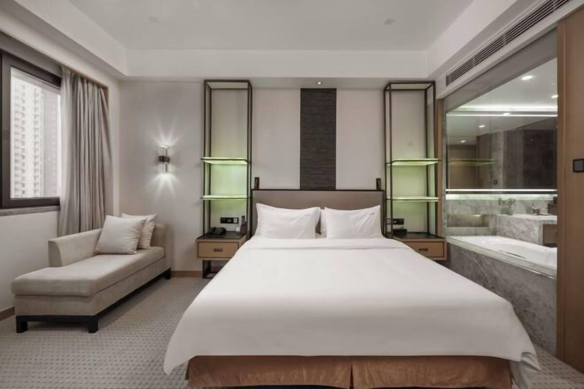 A hotel room with a large bed, a sofa, a nightstand, a lamp, and a bathroom with a bathtub and mirror.