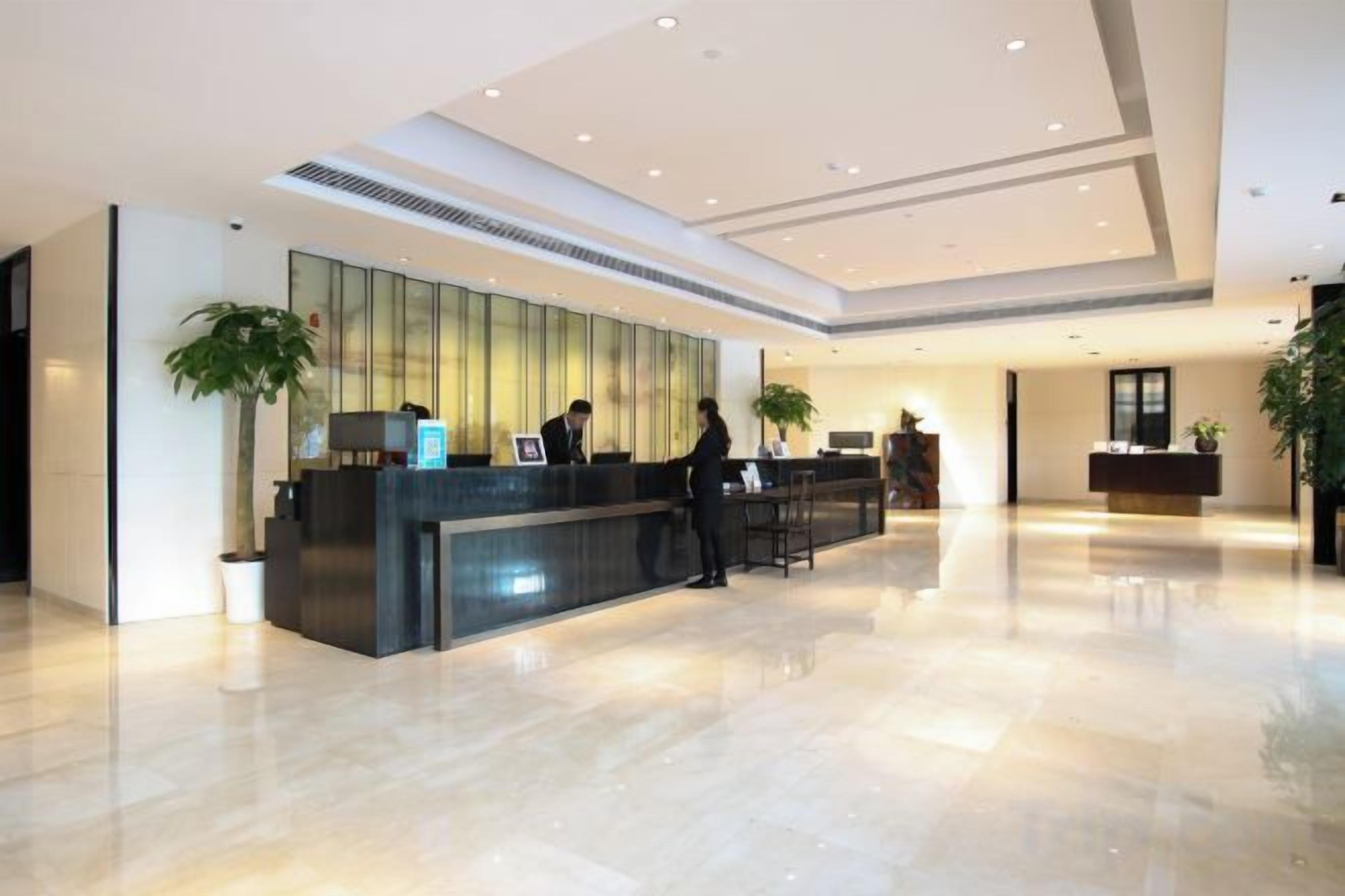 A hotel reception area with a reception desk, two staff members, and a large potted plant.